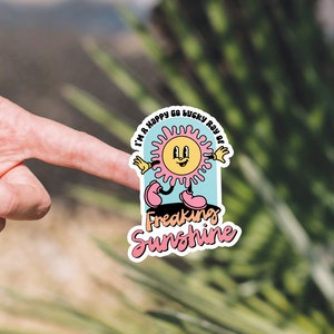 Op de afbeelding: Een sticker met een cartoonzon in roze en gele kleuren. De zon lacht en heeft een vrolijke uitdrukking. De tekst op de sticker luidt "I'm a happy go lucky ray of freaking sunshine".