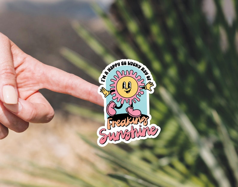 Op de afbeelding: Een sticker met een cartoonzon in roze en gele kleuren. De zon lacht en heeft een vrolijke uitdrukking. De tekst op de sticker luidt "I'm a happy go lucky ray of freaking sunshine".