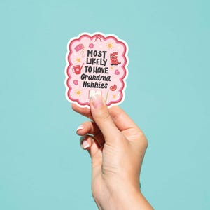 Może przedstawiać: R&oacute;żowa naklejka z napisem "MOST LIKELY TO HAVE Grandma Hobbies" w zabawnej czcionce. Naklejka ma falisty brzeg i zawiera małe ilustracje przedmiot&oacute;w rzemieślniczych. Trzymana w dłoni na turkusowym tle.