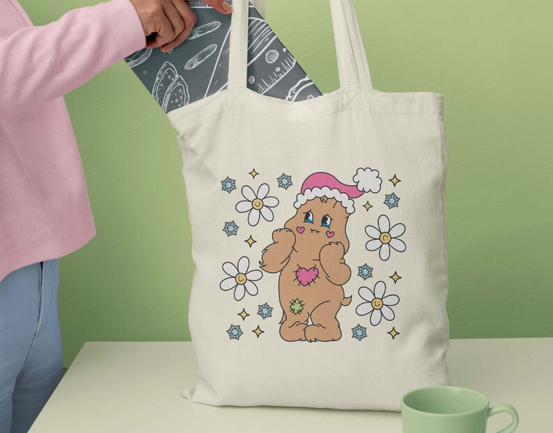 May include: A white tote bag featuring a brown bear wearing a pink Santa hat and surrounded by colourful flowers. The bear is holding a pink heart. The bag has a white background with a floral design.