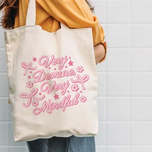 May include: A white canvas tote bag with pink text that reads "Very Demure, Very Mindful". The text is surrounded by pink stars and flowers.