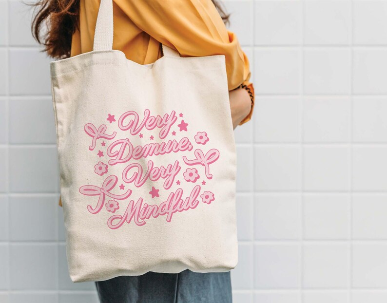 May include: A white canvas tote bag with pink text that reads "Very Demure, Very Mindful". The text is surrounded by pink stars and flowers.