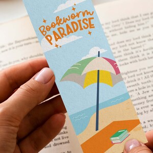 May include: A colorful bookmark with the text "Bookworm Paradise" features a beach scene with an umbrella, ocean, and a stack of books. The bookmark is held in a person's hands, with a book in the background.