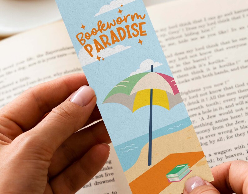 May include: A colorful bookmark with the text "Bookworm Paradise" features a beach scene with an umbrella, ocean, and a stack of books. The bookmark is held in a person's hands, with a book in the background.