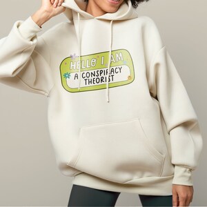 May include: A cream-colored hoodie with a green name tag that says "Hello I am a conspiracy theorist" in a colorful font.
