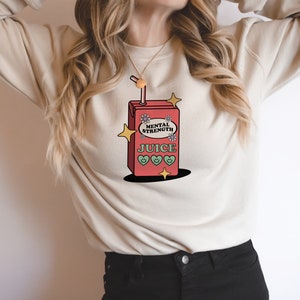 Puede incluir: Una sudadera blanca con un gr&aacute;fico de una caja de jugo roja con el texto "Mental Strength Juice" y corazones verdes. La caja de jugo tiene una pajita que sobresale.