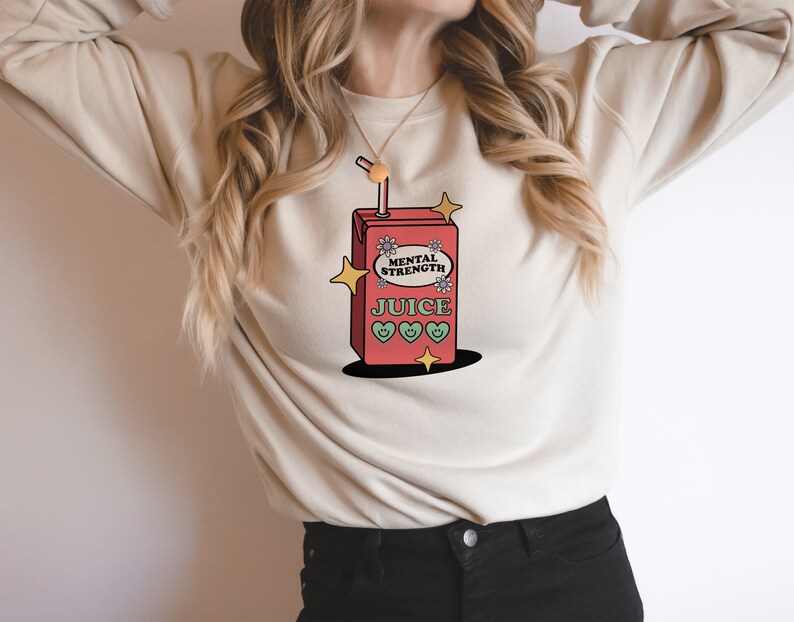 Puede incluir: Una sudadera blanca con un gr&aacute;fico de una caja de jugo roja con el texto "Mental Strength Juice" y corazones verdes. La caja de jugo tiene una pajita que sobresale.