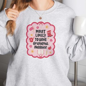 Może przedstawiać: Jasnoszara bluza z r&oacute;żowo-czerwoną grafiką z napisem "MOST LIKELY TO HAVE Grandma Hobbies". Grafika zawiera ilustracje konewki, but&oacute;w i książki. Trzymany jest biały kubek.