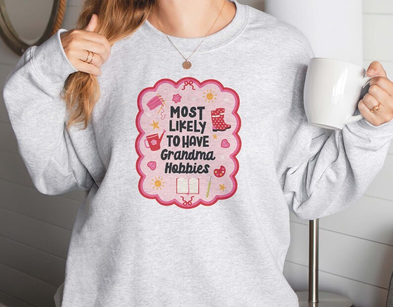 Może przedstawiać: Jasnoszara bluza z r&oacute;żowo-czerwoną grafiką z napisem "MOST LIKELY TO HAVE Grandma Hobbies". Grafika zawiera ilustracje konewki, but&oacute;w i książki. Trzymany jest biały kubek.