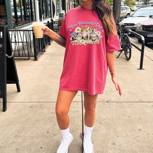 Op de afbeelding: Een vrouw draagt een rood oversized T-shirt met een afbeelding van kittens en de tekst "You Passed The Vibe Check". Ze houdt een koffiebeker mee naar huis vast en draagt witte sneakers.