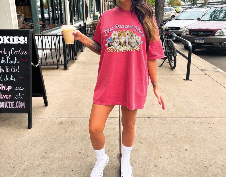Op de afbeelding: Een vrouw draagt een rood oversized T-shirt met een afbeelding van kittens en de tekst "You Passed The Vibe Check". Ze houdt een koffiebeker mee naar huis vast en draagt witte sneakers.