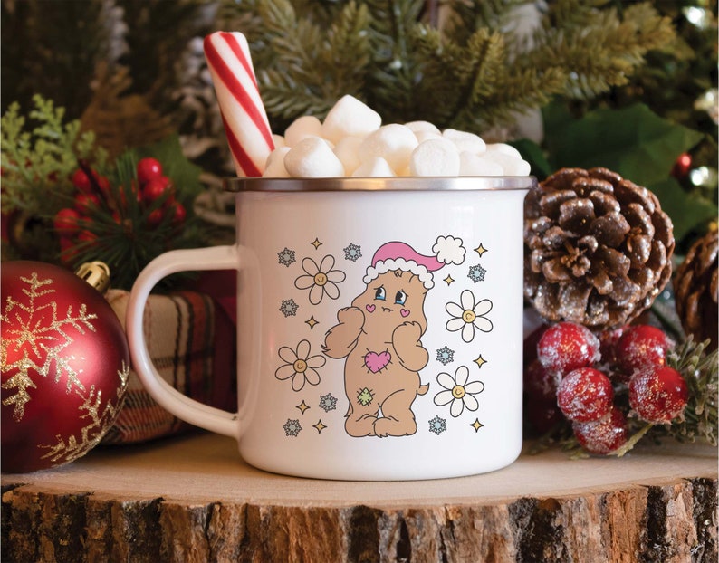 May include: White enamel mug featuring a brown bear wearing a pink Santa hat and surrounded by white daisies. The mug is filled with marshmallows and a red and white candy cane.