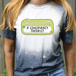 May include: A white t-shirt with a yellow and green name tag that says "Hello I am a conspiracy theorist". The t-shirt has a black and white splatter pattern.