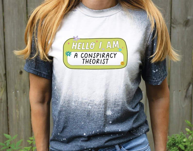 May include: A white t-shirt with a yellow and green name tag that says "Hello I am a conspiracy theorist". The t-shirt has a black and white splatter pattern.