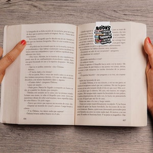 May include: A person's hands are holding open a book with a bookmark that says "Books are cheaper therapy,  much more effective". The book is open to a page with text in Spanish.