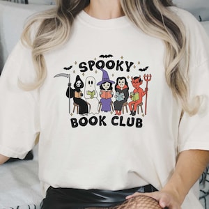 Puede incluir: Camiseta color crema con un dise&ntilde;o de "Spooky Book Club". El dise&ntilde;o incluye figuras de dibujos animados de la Parca, un fantasma, una bruja, un vampiro y un diablo, todos leyendo libros. Murci&eacute;lagos y estrellas rodean el texto.