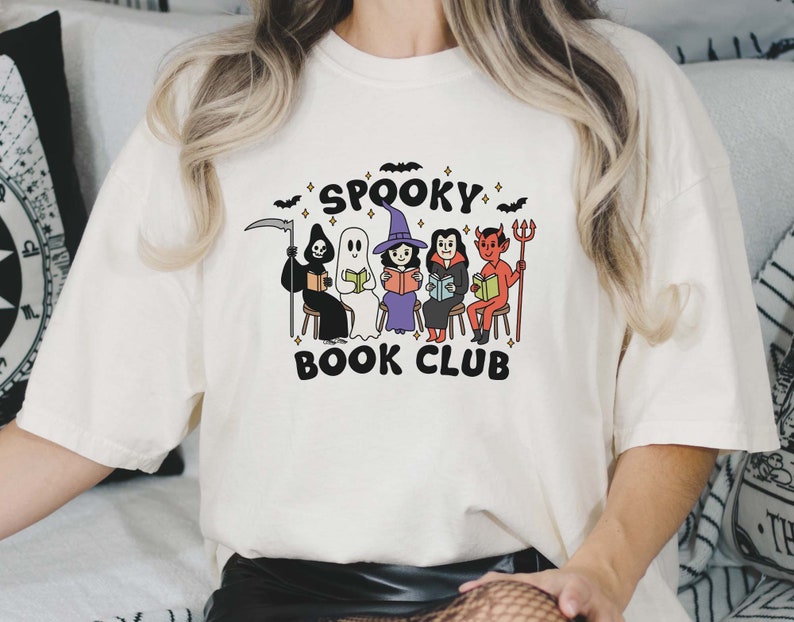 Puede incluir: Camiseta color crema con un dise&ntilde;o de "Spooky Book Club". El dise&ntilde;o incluye figuras de dibujos animados de la Parca, un fantasma, una bruja, un vampiro y un diablo, todos leyendo libros. Murci&eacute;lagos y estrellas rodean el texto.