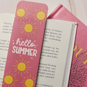 May include: Pink bookmark with yellow smiling suns and the words "hello SUMMER" in white. The bookmark is inserted into a book with white pages and a pink cover.