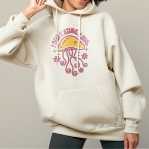 K&ouml;nnte beinhalten: Ein cremefarbener Hoodie mit einem Aufdruck einer rosa Qualle und dem Text "Ich steche nicht, ich umarme".