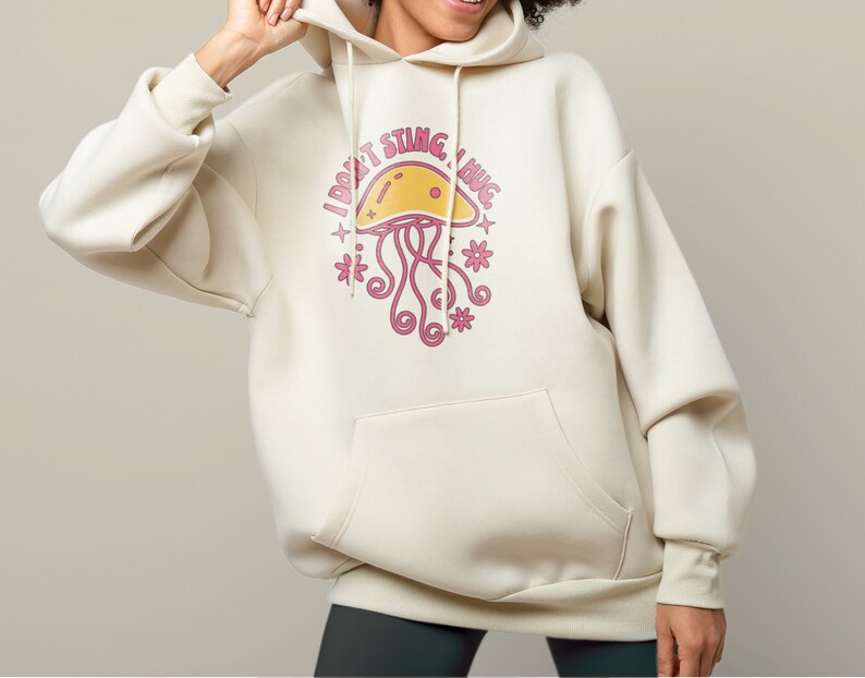 K&ouml;nnte beinhalten: Ein cremefarbener Hoodie mit einem Aufdruck einer rosa Qualle und dem Text "Ich steche nicht, ich umarme".