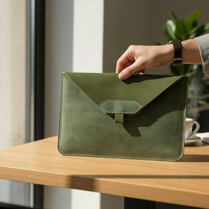 Puede incluir: Funda para portátil de cuero verde oliva con cierre de solapa y una pequeña correa. El diseño es simple y moderno, adecuado para llevar un portátil o una tableta. Sobre una mesa de madera.