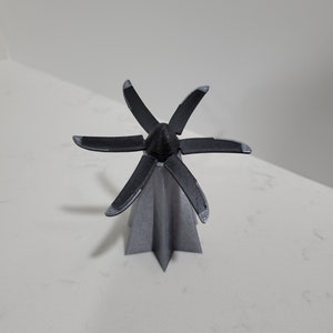 Peut inclure: Un modèle imprimé en 3D d'une petite base en forme d'étoile grise avec une hélice noire à cinq pales sur le dessus.