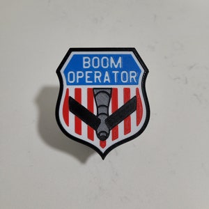 May include: A black and white patch with a red, white, and blue striped background. The patch has a gray silhouette of a plane with the text "BOOM OPERATOR" in white letters.