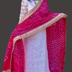 May include: A vibrant magenta dupatta with a gold trim and white dotted pattern. The dupatta is draped over a white embroidered outfit. The outfit has long sleeves with a lace design. The background is a solid dark gray.