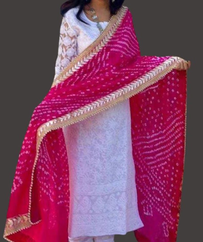 May include: A vibrant magenta dupatta with a gold trim and white dotted pattern. The dupatta is draped over a white embroidered outfit. The outfit has long sleeves with a lace design. The background is a solid dark gray.