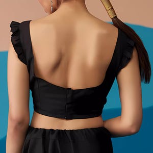 May include: A black crop top with frilled cap sleeves and a square neckline. The back of the top features a fitted design with a central seam and a subtle flare at the waist. The top is paired with a matching black skirt.