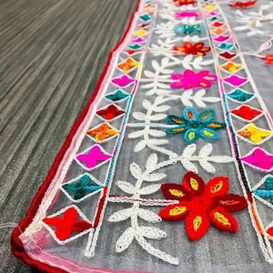 May include: White sheer fabric with colourful embroidered flowers and geometric patterns. The fabric has a red border.