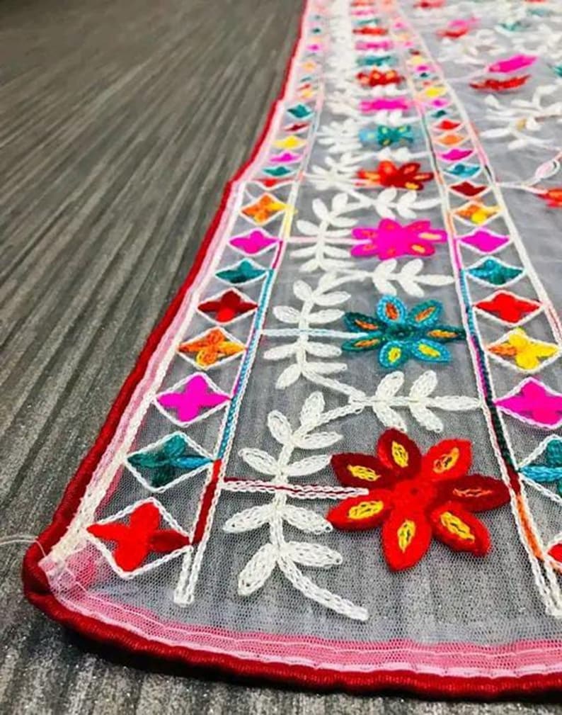 May include: White sheer fabric with colourful embroidered flowers and geometric patterns. The fabric has a red border.
