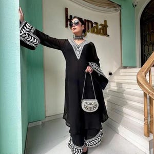 May include: Black ensemble featuring a long-sleeved top and matching pants, both adorned with white embroidered detailing. The top has a V-neck and flared sleeves. A small, silver-toned purse with a chain strap completes the outfit. The outfit is worn by a person.