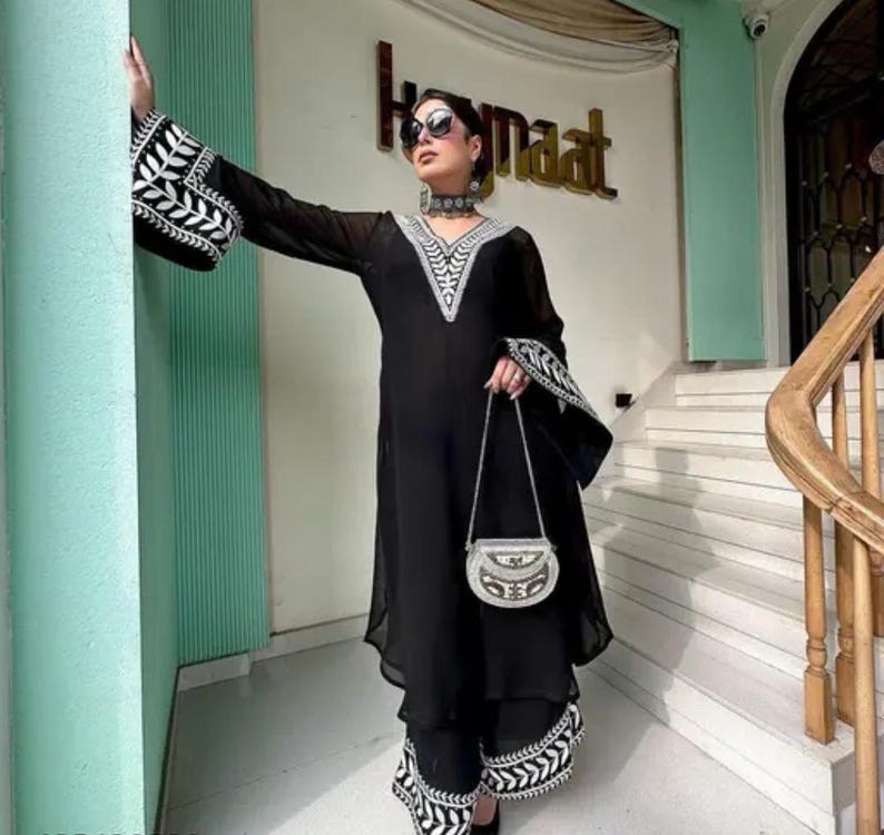 May include: Black ensemble featuring a long-sleeved top and matching pants, both adorned with white embroidered detailing. The top has a V-neck and flared sleeves. A small, silver-toned purse with a chain strap completes the outfit. The outfit is worn by a person.