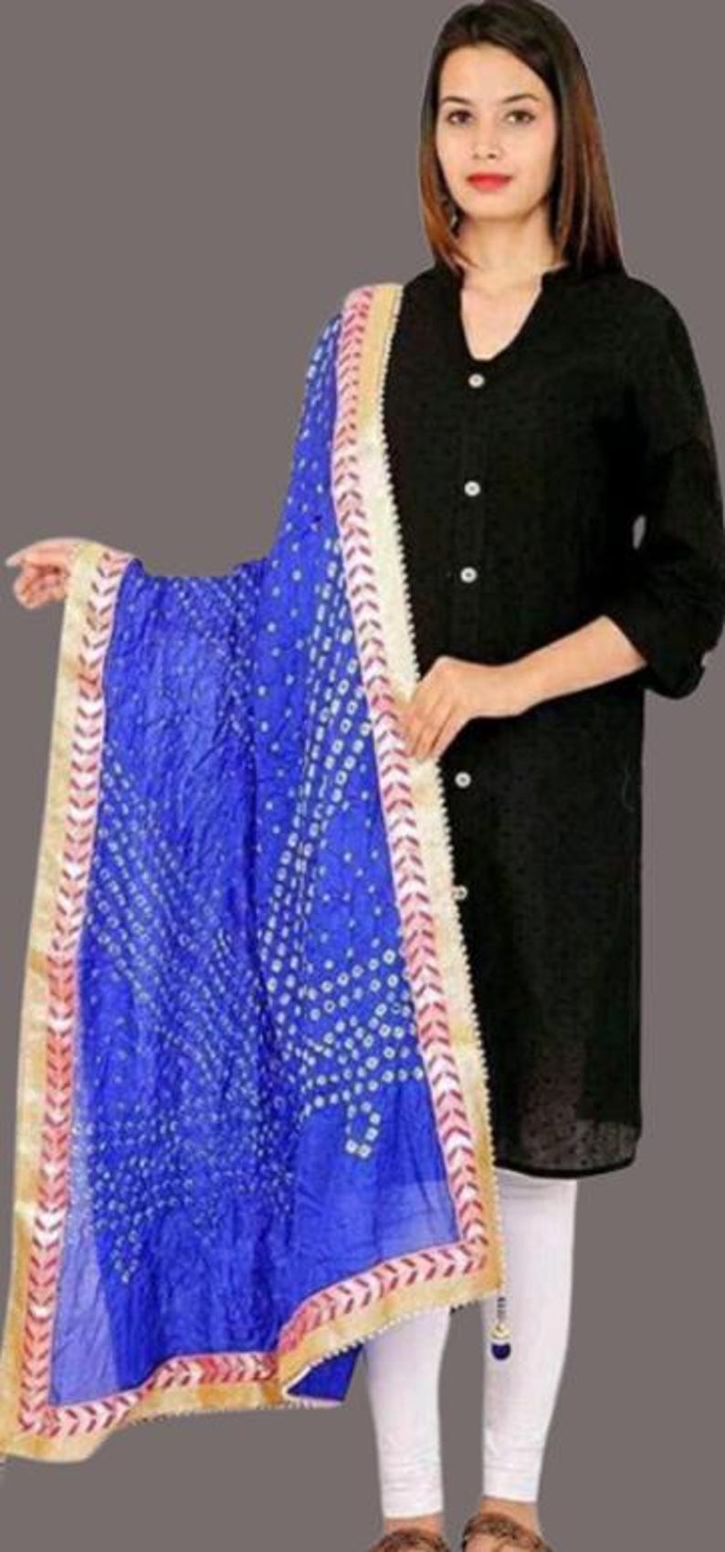 May include: A vibrant blue dupatta with a white dotted pattern and gold trim. The scarf is draped over a black top and white leggings. The dupatta's border has a pink and gold design.