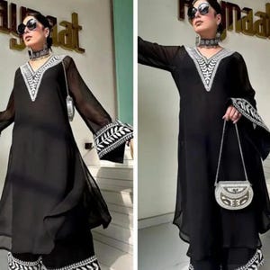 May include: A black ensemble featuring a long-sleeved tunic and wide-leg pants, both adorned with intricate white embroidery along the neckline, cuffs, and hem. A small, silver handbag complements the outfit. The model is wearing sunglasses and a matching choker.