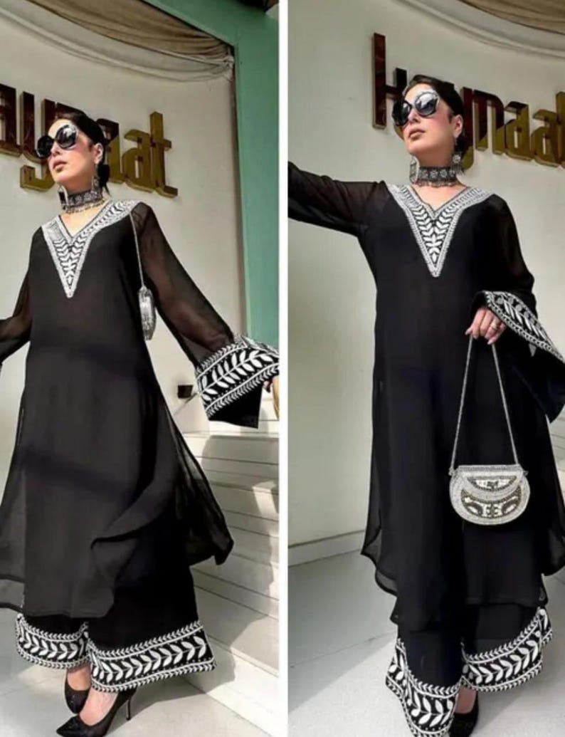 May include: A black ensemble featuring a long-sleeved tunic and wide-leg pants, both adorned with intricate white embroidery along the neckline, cuffs, and hem. A small, silver handbag complements the outfit. The model is wearing sunglasses and a matching choker.