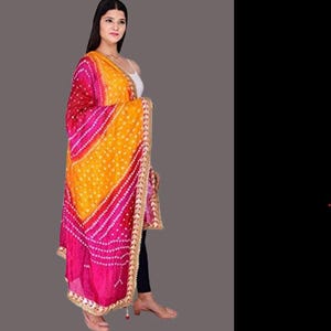 May include: A vibrant, patterned scarf in shades of red, pink, and yellow. The scarf features a detailed design with white dots and a gold-colored trim. The fabric is draped, showcasing the colors and pattern.