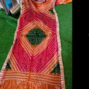 May include: A vibrant textile with a geometric pattern in shades of red, orange, and green. The fabric features a diamond-shaped design in the center, with white accents and a decorative border. The textile is likely a scarf or shawl.