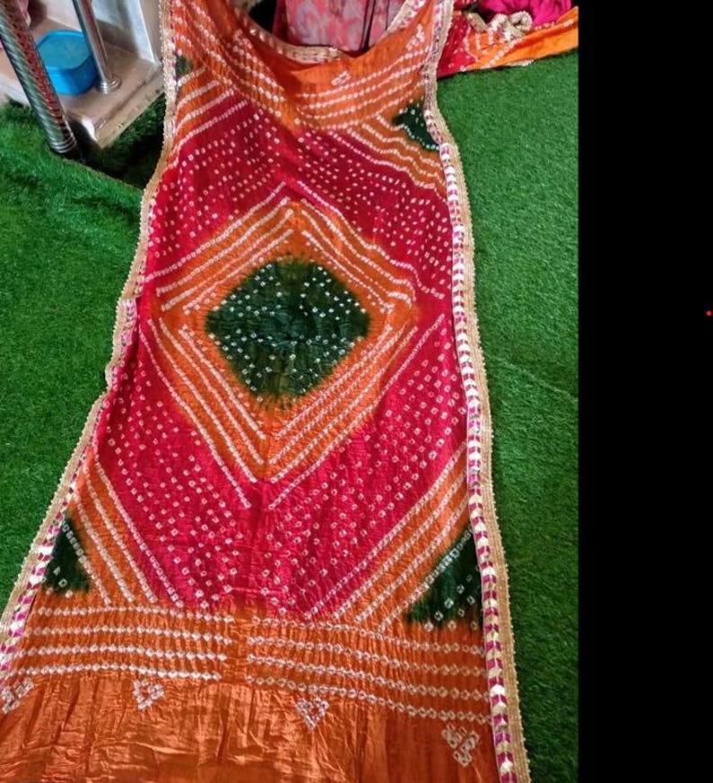 May include: A vibrant textile with a geometric pattern in shades of red, orange, and green. The fabric features a diamond-shaped design in the center, with white accents and a decorative border. The textile is likely a scarf or shawl.