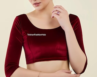 May include: A deep red velvet blouse with three-quarter length sleeves. The scoop neckline and fitted design create a classic silhouette. The text "TrishanFashionHub" is printed on the blouse. A bracelet and ring are also visible.