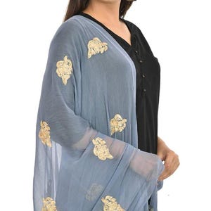 May include: A light blue sheer scarf with gold embroidered floral designs. The scarf is draped over a person wearing a black top.