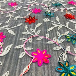 May include: White sheer fabric with embroidered floral designs in various colours, including pink, red, blue, and yellow. The flowers are arranged in a repeating pattern on the fabric.