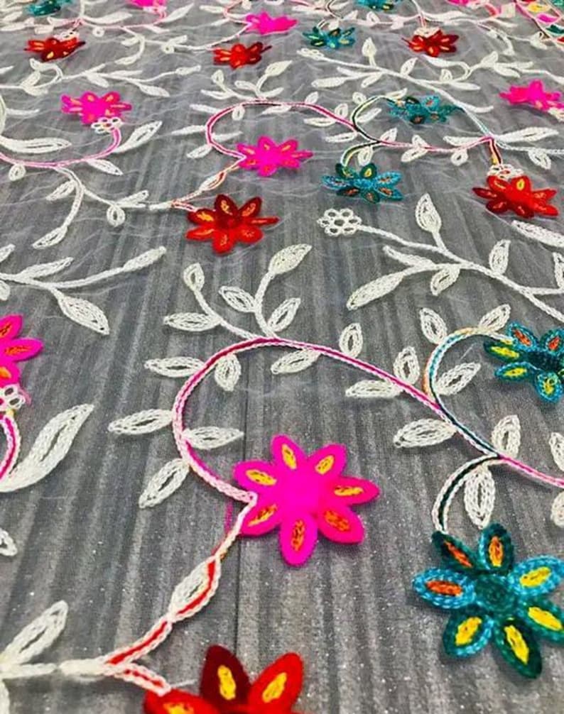 May include: White sheer fabric with embroidered floral designs in various colours, including pink, red, blue, and yellow. The flowers are arranged in a repeating pattern on the fabric.