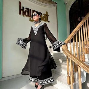 May include: A black ensemble featuring a long-sleeved tunic and matching pants. The tunic has a V-neck and flared sleeves with white embroidered details. The pants also have white embroidery at the hem. The outfit is worn with sunglasses and a choker.