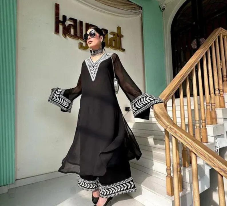 May include: A black ensemble featuring a long-sleeved tunic and matching pants. The tunic has a V-neck and flared sleeves with white embroidered details. The pants also have white embroidery at the hem. The outfit is worn with sunglasses and a choker.