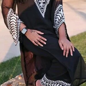 May include: Black two-piece outfit with white embroidered leaf patterns. The top has a V-neck and long sleeves. The pants have matching embroidery at the hem. The outfit is worn with black sandals.