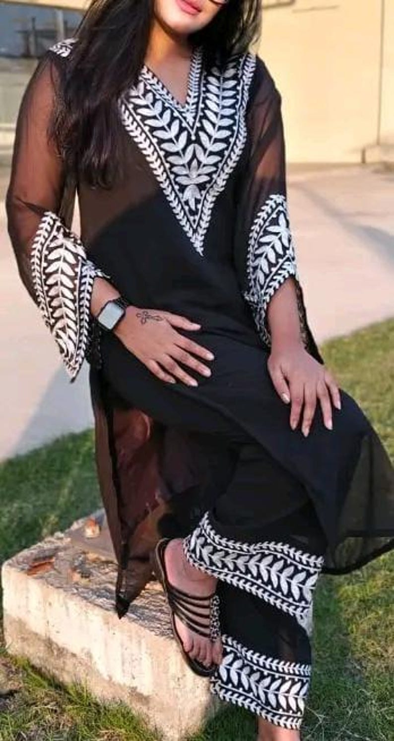 May include: Black two-piece outfit with white embroidered leaf patterns. The top has a V-neck and long sleeves. The pants have matching embroidery at the hem. The outfit is worn with black sandals.