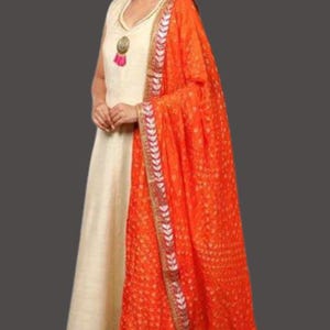 May include: A cream-colored, sleeveless dress with gold trim and a matching orange dupatta. The dupatta features a dotted pattern and decorative border. The dress has a V-neckline and a tassel pendant.