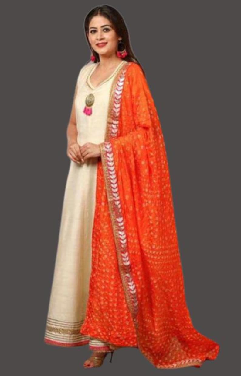 May include: A cream-colored, sleeveless dress with gold trim and a matching orange dupatta. The dupatta features a dotted pattern and decorative border. The dress has a V-neckline and a tassel pendant.